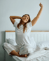 woman in pajamas sitting on a white bed, stretching with her arms raised after finding relief from sleep and energy problems