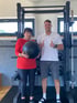 Eine Seniorin hält einen Medizinball neben einem Trainer vor einem Kraftgerät im Fitnessstudio.