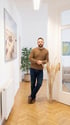Ein Mann mit Bart, braunem Pullover und Jeans steht lässig in einem hellen Flur mit Fischgrätparkett.