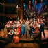 Große Gruppe von Schauspielern in Vintage-Kostümen auf einer Bühne, die jubeln und winken, im Hintergrund Stadtkulisse.