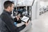 Techniker prüft Bauteil an einer Industriemaschine mithilfe eines Tablets in einer hellen Werkshalle.