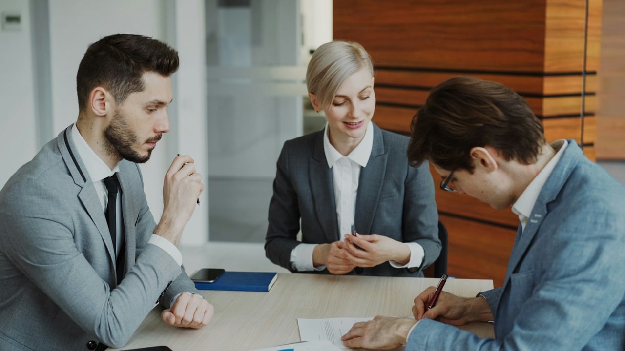 Drei Personen in Businesskleidung an einem Besprechungstisch, ein Mann unterschreibt ein Dokument