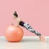 Frau in Sportkleidung macht einen Unterarmstütz auf einem großen orangefarbenen Gymnastikball vor rosa Wand.