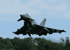 Dunkler Kampfjet hebt über Baumwipfeln mit Rauchspur in den blauen Himmel ab.