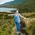A woman in a gray puffer jacket stands on a mountain trail with arms outstretched toward a lake and green hills.