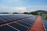 Solarpaneele auf einem roten Wellblechdach unter blauem Himmel mit Hügeln und Bäumen im Hintergrund.