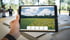 Hand hält ein Tablet mit einer Landschafts-App, die eine Wiese mit Blumen, Wald und blauem Himmel zeigt.