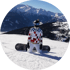 Snowboarder in weißer und rot-karierter Jacke, weißer Hose, Helm und Skibrille kniet im Schnee mit Bergpanorama im Hintergrund.
