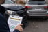 Mann hält Klemmbrett mit "KEINE KOSTEN", im Hintergrund zwei nach einem Unfall beschädigte Autos.