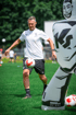 Ein Fußballspieler in Weiß und Schwarz jongliert einen Ball auf einem grünen Feld neben einer aufblasbaren Puppe.