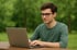 Junger Mann mit Brille tippt auf einem Laptop an einem Holztisch im Freien.