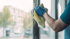 Eine Hand mit blauem Handschuh wischt mit einem gelben Tuch eine Fensterscheibe.
