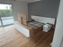 Schlafzimmer mit Holzboden, Bett und TV-Möbel in Holzoptik und Weiß vor grauer Wand, Balkontür links.