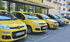 Fünf gelbe Autos mit Logos und Kennzeichen parken in einer Reihe vor einem Gebäude.