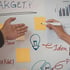 Hände schreiben auf ein Whiteboard mit Notizen, Glühbirnenzeichnung und Diagrammen.