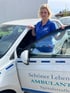 Blonde Frau in blauem Polo-Shirt mit Logo steht lächelnd an der geöffneten Tür eines weißen Autos.