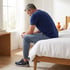 A man in a blue t-shirt and joggers sits on the edge of a bed in a bright bedroom, looking down with his hands clasped.