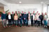 Eine Gruppe von siebzehn lächelnden Mitarbeitern in Business-Casual-Kleidung posiert für ein Teamfoto in einem hellen Büro.