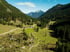 Grünes Tal mit Bauernhof und bewaldeten Bergen unter blauem Himmel.
