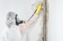 Eine Person in einem Schutzanzug, Gasmaske und gelben Handschuhen reinigt schwarzen Schimmel von einer Wand.