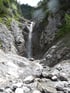 Ein Wasserfall stürzt über graue Felsen in ein felsiges Bachbett mit Geröll und einigen grünen Pflanzen.