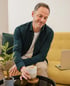 Ein lächelnder Mann sitzt auf einem gelben Sofa, hält eine Kaffeetasse und schaut auf einen Laptop.