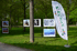 Ein weißes Banner mit "KRÜN e.V." und "www.kunst-gruen.de" steht neben drei Rahmen mit Gemälden auf einer Wiese.
