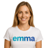 Lächelnde Frau mit langen blonden Haaren trägt ein weißes T-Shirt mit dem blauen Schriftzug emma vor schwarzem Hintergrund.