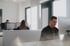 Ein Mann mit Brille und Kopfhörern arbeitet am Computer in einem Büro mit mehreren Arbeitsplätzen.