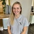 Eine lächelnde junge Frau mit blonden Haaren trägt ein graues Poloshirt mit dem Logo "Fitness for Travel".