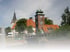 Kirche mit spitzem Turm und daneben stehendes Backsteingebäude mit grünem Dach und Ausleger über Wasser.