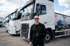 Ein Mann mit Sonnenbrille und schwarzer Jacke steht vor einer Reihe weißer Volvo-Lastwagen.