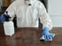 Person in Schutzanzug, Maske und blauen Handschuhen reinigt Tisch mit Sprühflasche und Tuch.