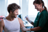 A medical professional in green scrubs gives a testosterone shot to a smiling man in a white tank top.