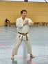 Keigo Shimizu Sensei in weißem Karate-Gi und braunem Gürtel demonstriert eine Handhaltung in einer Turnhalle.