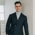 Lächelnder Mann im dunkelblauen Zweireiher-Sakko und grauem Rollkragenpullover lehnt an einer hellen Wand.