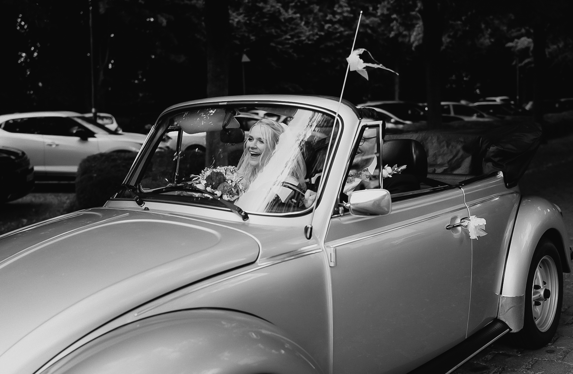 Schwarz-Weiß-Aufnahme: Die Braut sitzt im offenen VW-Käfer-Cabrio am Gut Dyckhof in Meerbusch.