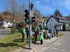 Wandergruppe wartet an einer Bushaltestelle neben einer grünen Ampel in einer dörflichen Straße bei Sonnenschein.