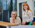 Zwei lächelnde Frauen, eine sitzt am Schreibtisch mit Monitor, die andere steht daneben, vor einem Wandbild mit Bäumen.