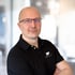 Mann mit Glatze und Brille, der ein schwarzes Poloshirt mit "S PIXELS"-Logo trägt, verschränkt die Arme.