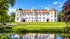 Weißes Schloss Celle mit roten Dächern hinter einer grünen Wiese und einem Teich unter blauem Himmel.