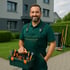 Ein lächelnder Mann in grüner Arbeitskleidung hält einen Werkzeugkasten vor einem Haus mit Rasen.