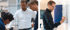 Ein Mann in weißem Hemd spricht mit zwei jüngeren Männern; ein Mann in schwarzem Hemd schreibt auf eine Tafel.