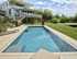Moderner Pool im Garten eines Hauses mit Terrassen und grüner Wiese unter blauem Himmel.