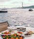 Table set with food and wine glasses on a boat, with the Bosphorus Bridge and boats in the background.