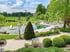 Moderner Schwimmteich mit Sonnendeck, Liegestühlen und gepflegter Gartenanlage mit Buchsbäumen und Skulpturen unter blauem Himmel.