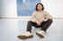 James Lanigan smiling with dark hair and a beard sits on a light floor, wearing a light sweater and dark jeans.
