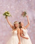 Zwei Bräute in weißen Hochzeitskleidern halten jubelnd ihre Blumensträuße vor einem rosa Bokeh-Hintergrund hoch.