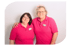 Zwei lächelnde Frauen in pinkfarbenen Poloshirts und Brillen, eine den Arm um die andere gelegt.