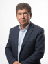 Headshot of a man with short, dark, curly hair, wearing a dark blue suit jacket and light blue shirt.